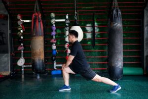 An Asian male athlete performs a lunge stretch in a training gym. The background shows various punching bags and fitness equipment, ready for an intense workout.