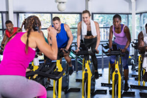 Fitness instructor leading diverse spin class participants pedaling on yellow bikes in gym studio.
