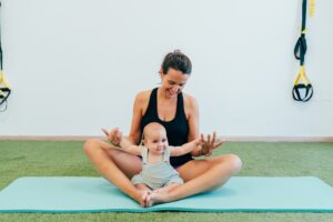 Mom practicing yoga with her baby sitting on her legs, on a fitness mat, in a gym.
